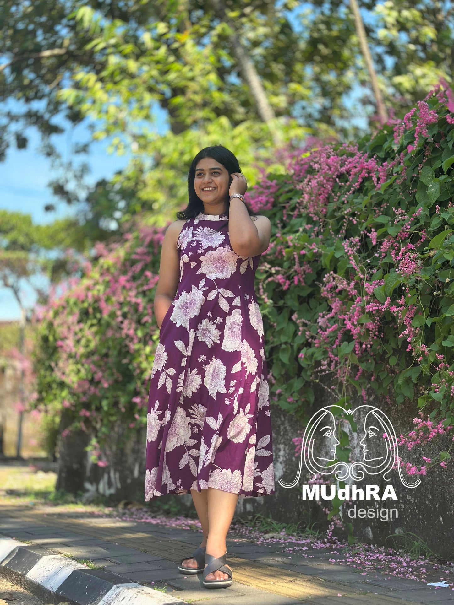 Woman in a purple floral dress standing in a garden with pink flowers and greenery.