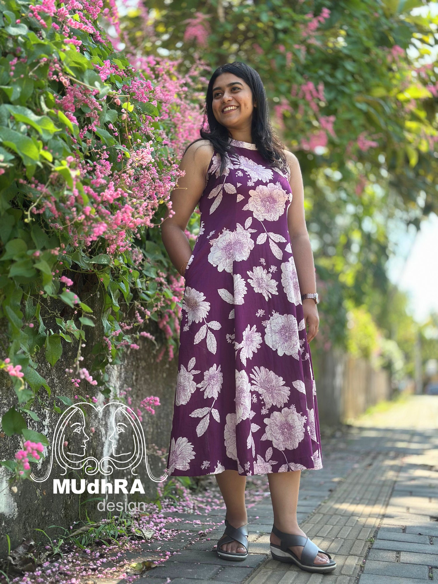 Woman in a purple floral dress standing next to pink flowers with 'Mudhra Design' logo.