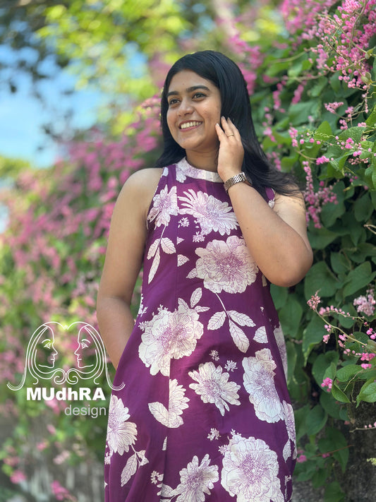 Woman in a purple floral dress standing in front of pink flowers, with 'MudhRA design' logo.